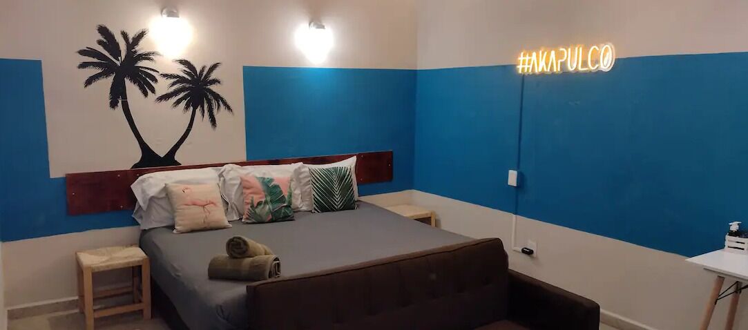 A bedroom with a bed, bedside tables, a wall with a palm tree mural, and a neon sign.