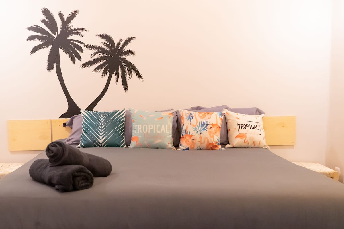 Ein Bett mit grauer Bettdecke, ein Kopfkissen mit tropischem Muster und ein weiteres Kopfkissen mit der Aufschrift "TROPICAL" und "TROPICAL".