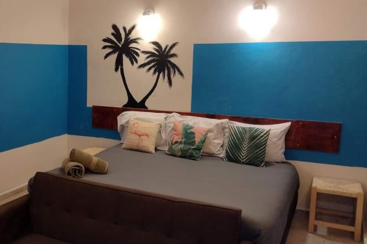 Ein Schlafzimmer mit einem Bett, Kissen, einem kleinen Hocker und einer Wand mit Palmenmotiv.