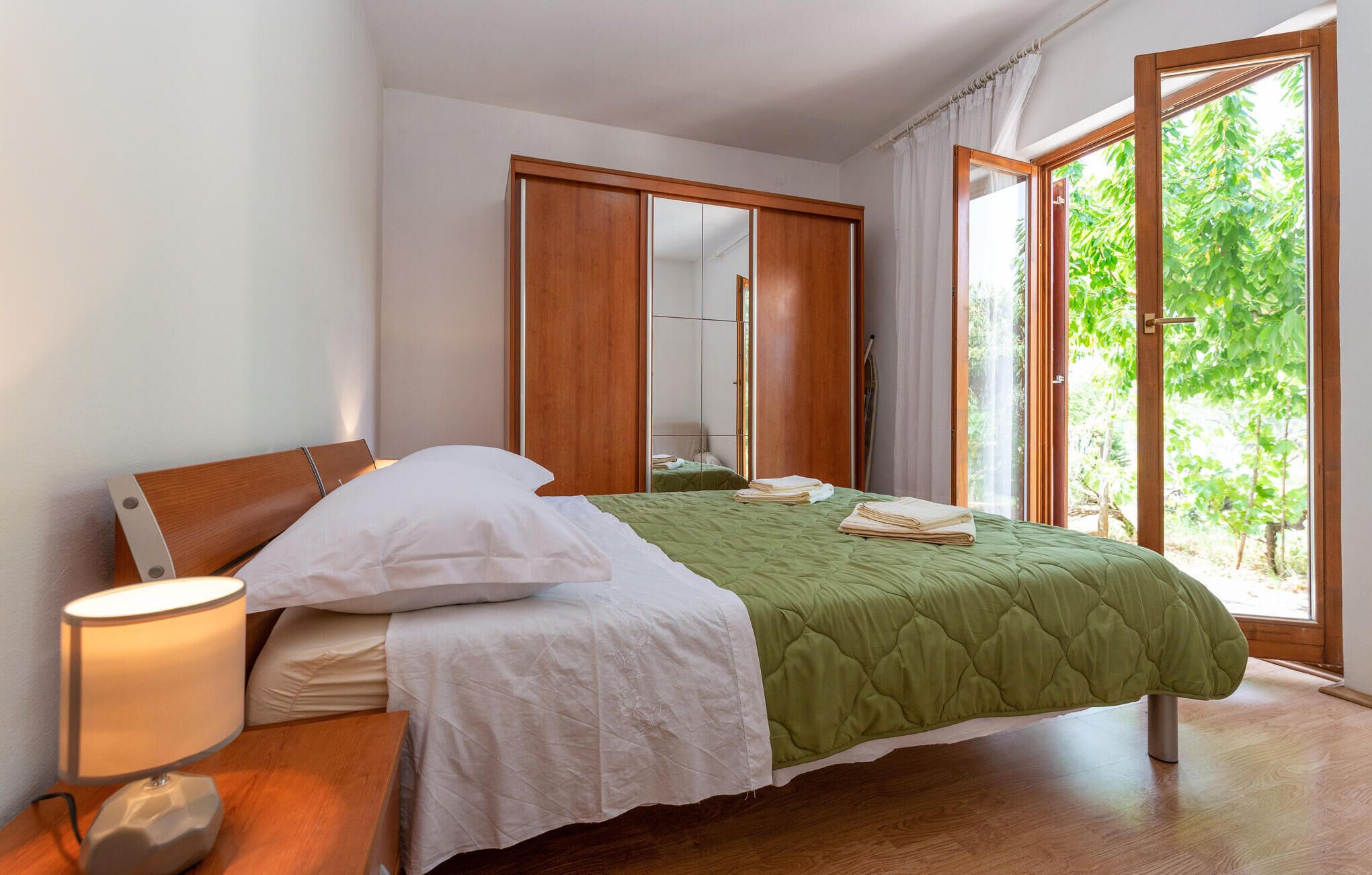 A bedroom with a bed, a bedside lamp, a wardrobe, and a window with a view of greenery.