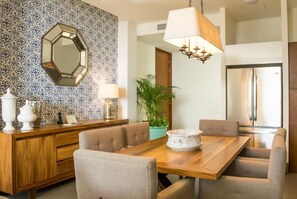 Uma sala de jantar com mesa de madeira, cadeiras estofadas bege, um lustre, um vaso com planta e um espelho decorativo na parede.