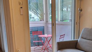 Una habitación con un sofá gris, una mesa con una revista, una mesita de madera y vistas a un patio con sillas y una mesa rojas.