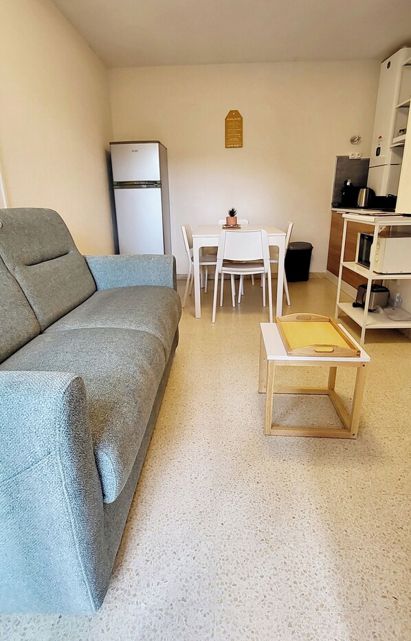 Ein kompakter Wohnraum mit Sofa, kleinem Tisch, Esstisch mit Stühlen, Kühlschrank und Küchenzeile mit Regalen und Geräten.