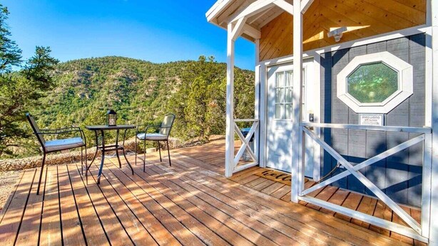 Una terraza de madera con muebles exteriores, un ventanal hexagonal y vistas a una ladera boscosa.