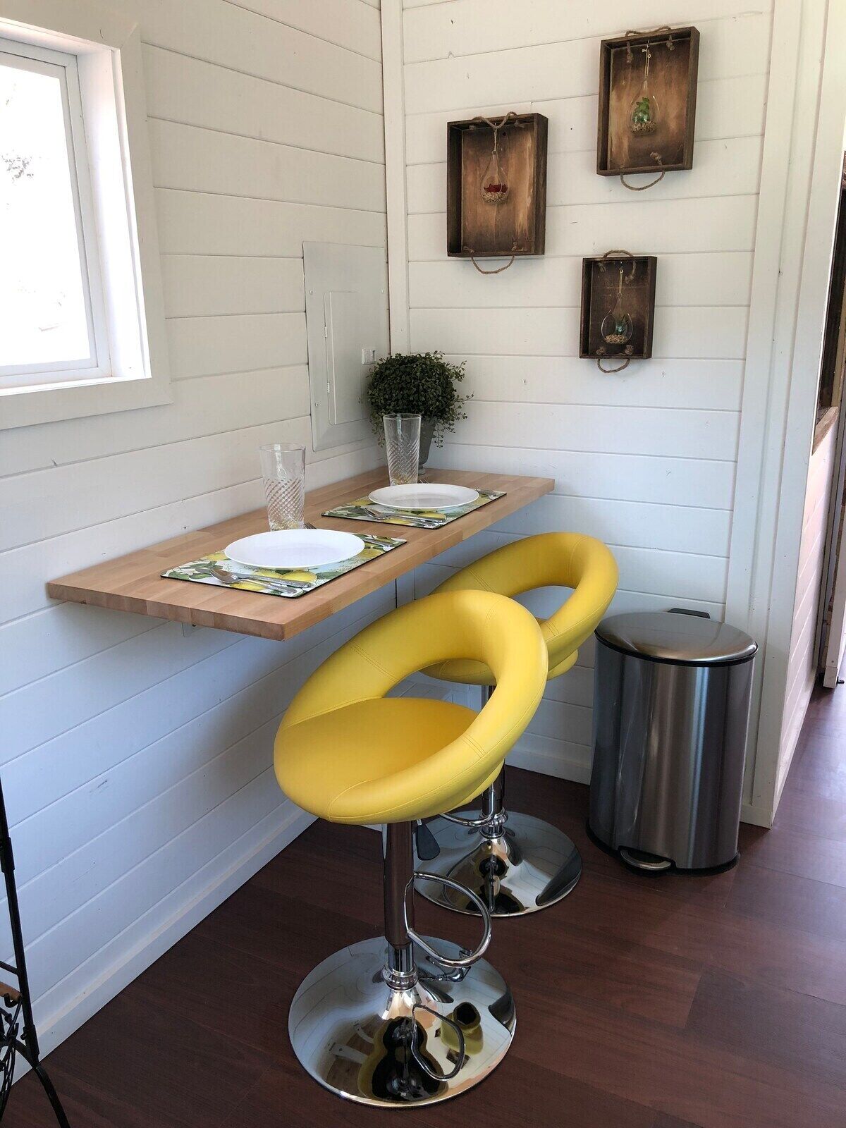 Une cuisine de taille réduite avec une table en bois, deux tabourets de bar jaunes, une poubelle et des éléments décoratifs muraux.