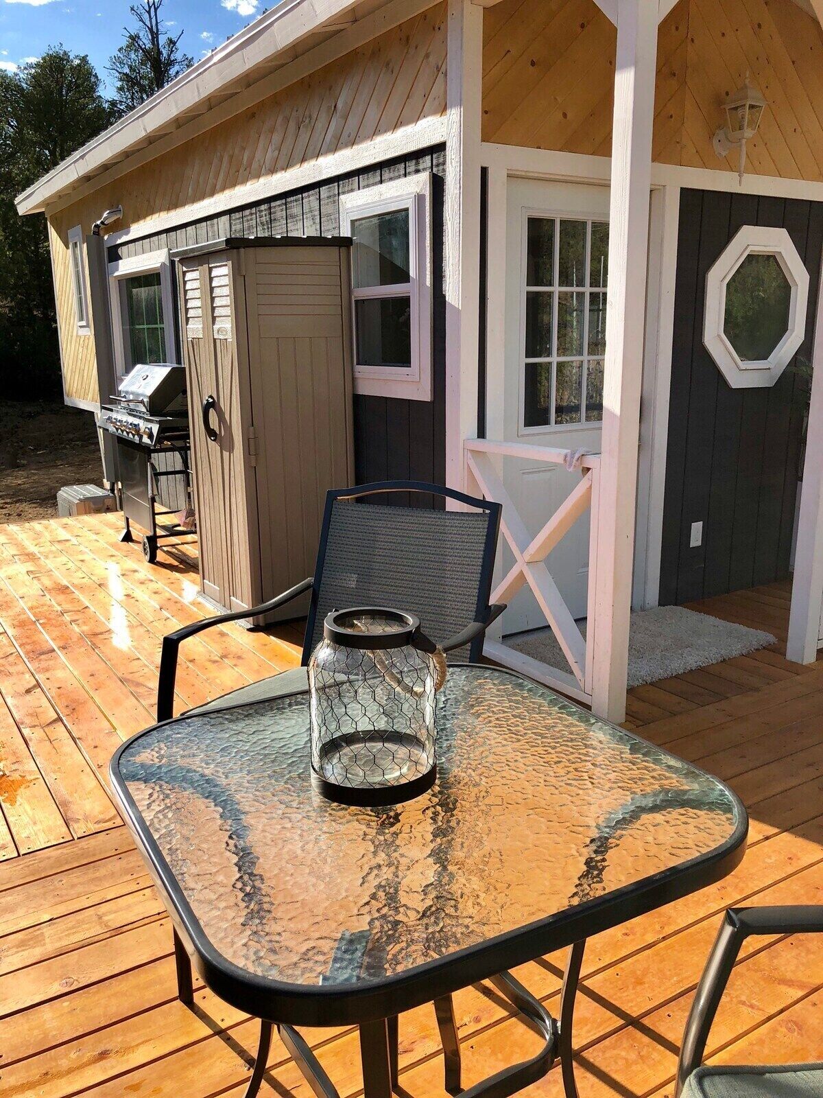A wooden deck with a glass table and chairs, a grill, and a covered porch area.