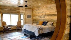A wooden cabin-style bedroom with a bed, two chairs, a ceiling fan, and a large window. - New! Juniper Ridge Luxury Lodge @ East Zion & Bryce (Orderville)