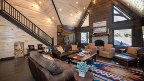 A spacious living room with a high ceiling, wooden walls, a fireplace, and a large window providing a view of the outdoors. - New! Juniper Ridge Luxury Lodge @ East Zion & Bryce (Orderville)
