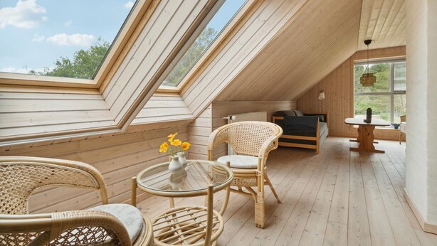 Een knus zolderkamertje met houten vloeren, een glazen tafel, rieten stoelen en een bed.
