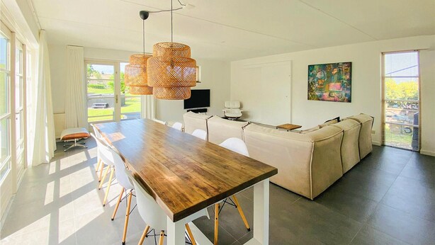 A modern kitchen with a long wooden table, white chairs, a beige sofa, and a TV.