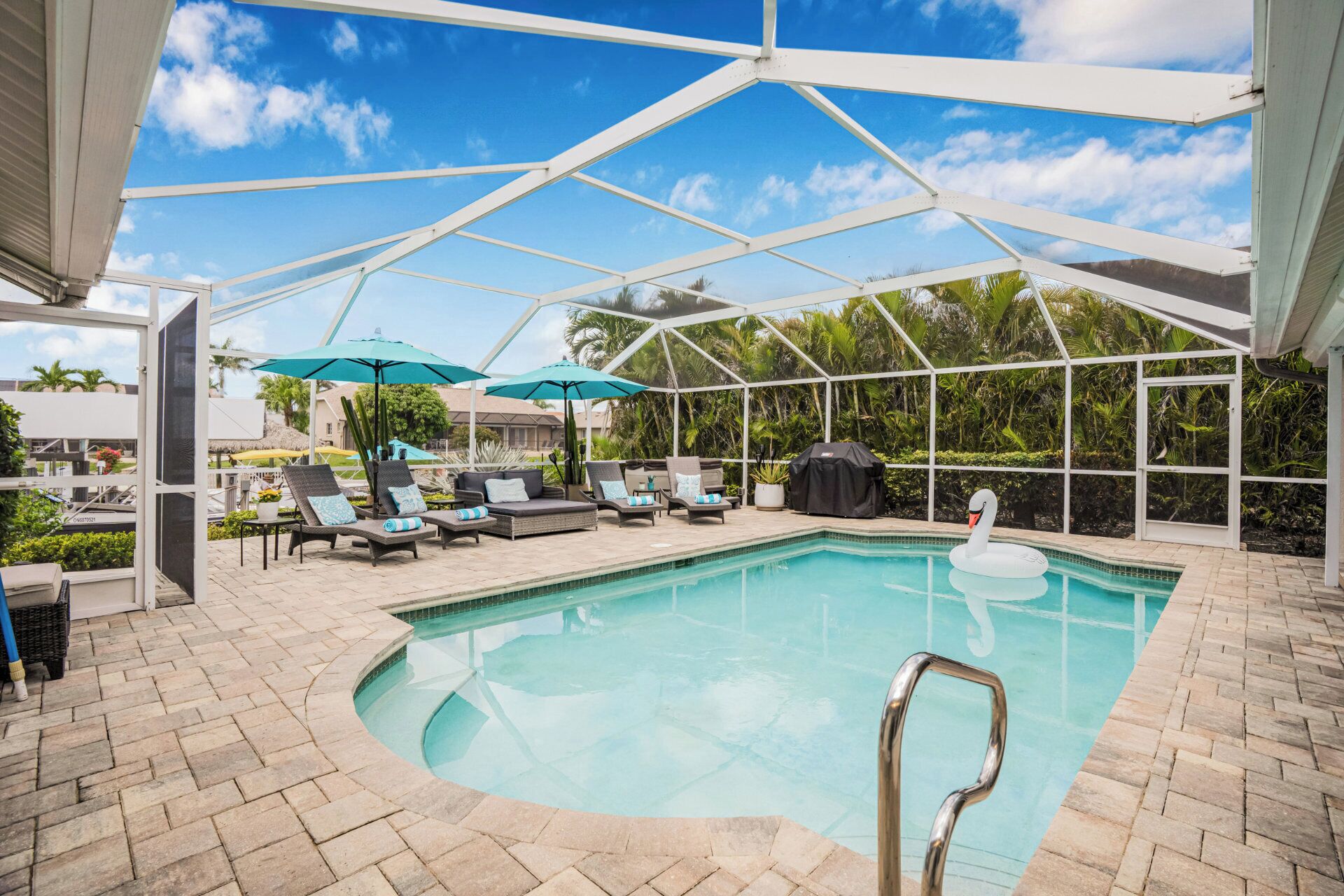A covered swimming pool with lounge chairs and umbrellas, surrounded by palm trees and a clear sky.