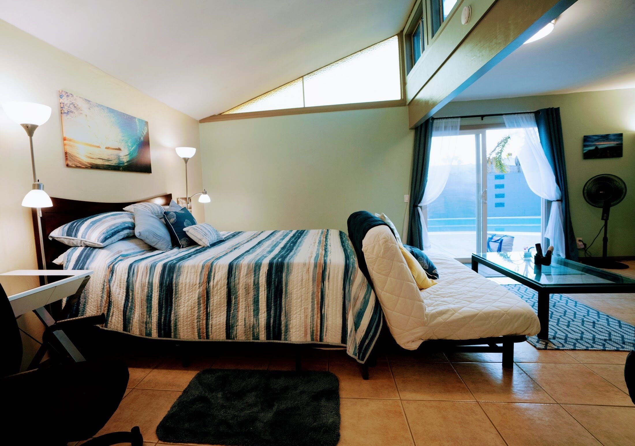 A hotel room with a bed, a desk, a chair, a black rug, a painting on the wall, and a window with curtains.