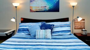 Ein Bett mit einer gestreiften, blau-weißen Tagesdecke, zwei Sternpilows und einem Wellenbild darüber. Auf jeder Seite des Bettes stehen zwei Nachttischlampen.