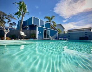 Een modern huis met een zwembad, palmbomen en een helderblauwe lucht.
