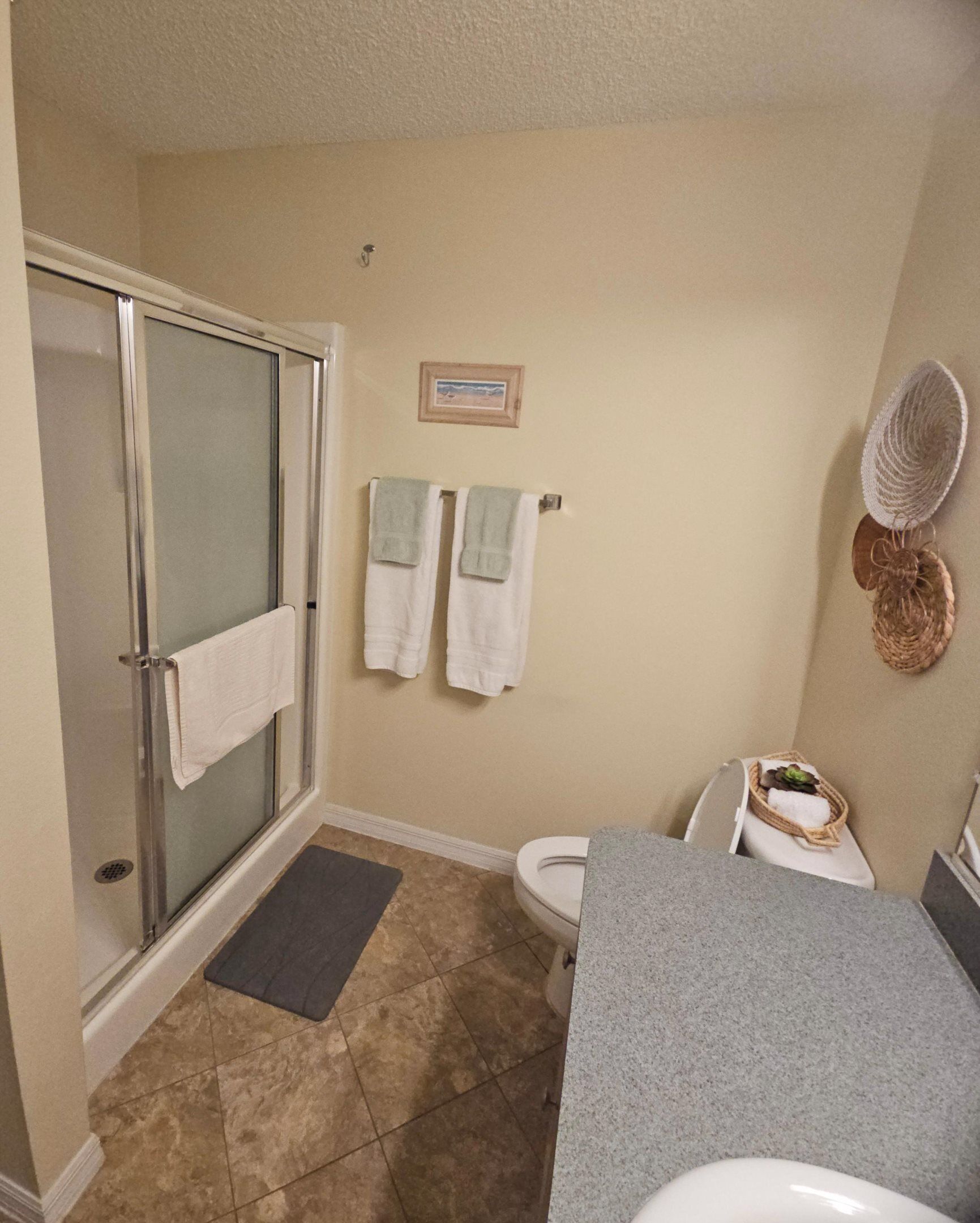 A bathroom with a glass shower enclosure, two white towels on a wall-mounted rack, a framed picture, a wicker basket with a towel, and a granite countertop.