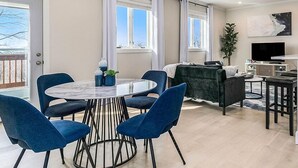 A modern living room with a round dining table, blue chairs, a dark green sofa, a flat-screen TV, and a large window with curtains.