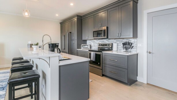 A modern kitchen with grey cabinets, a central island, stainless steel appliances, and a white backsplash.