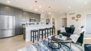 A modern kitchen with stainless steel appliances, a central island with bar stools, and a dining area with a round table and blue patterned rug.