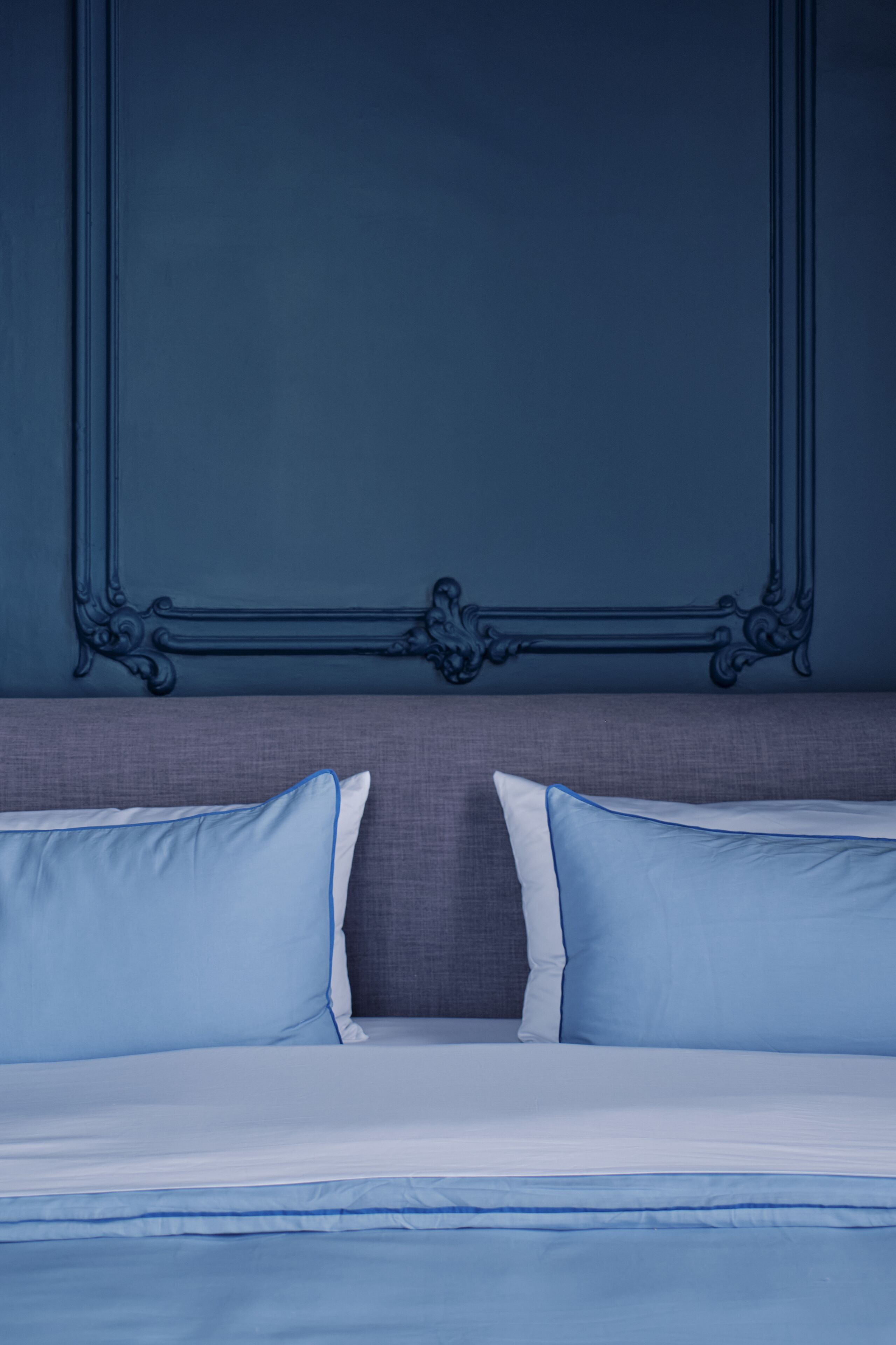 A bed with blue bedding and pillows, a grey headboard, and a dark blue wall with an ornate mirror.
