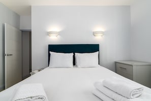 A neatly made bed with white linens and pillows, two bedside lamps, a nightstand, and a folded towel on the floor.