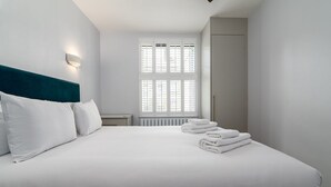 Un letto rifatto con lenzuola e cuscini bianchi, un comodino, una lampada a parete e una finestra con persiane.