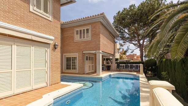 A swimming pool with a tiled deck, a brick house with shutters, and a palm tree in the background.