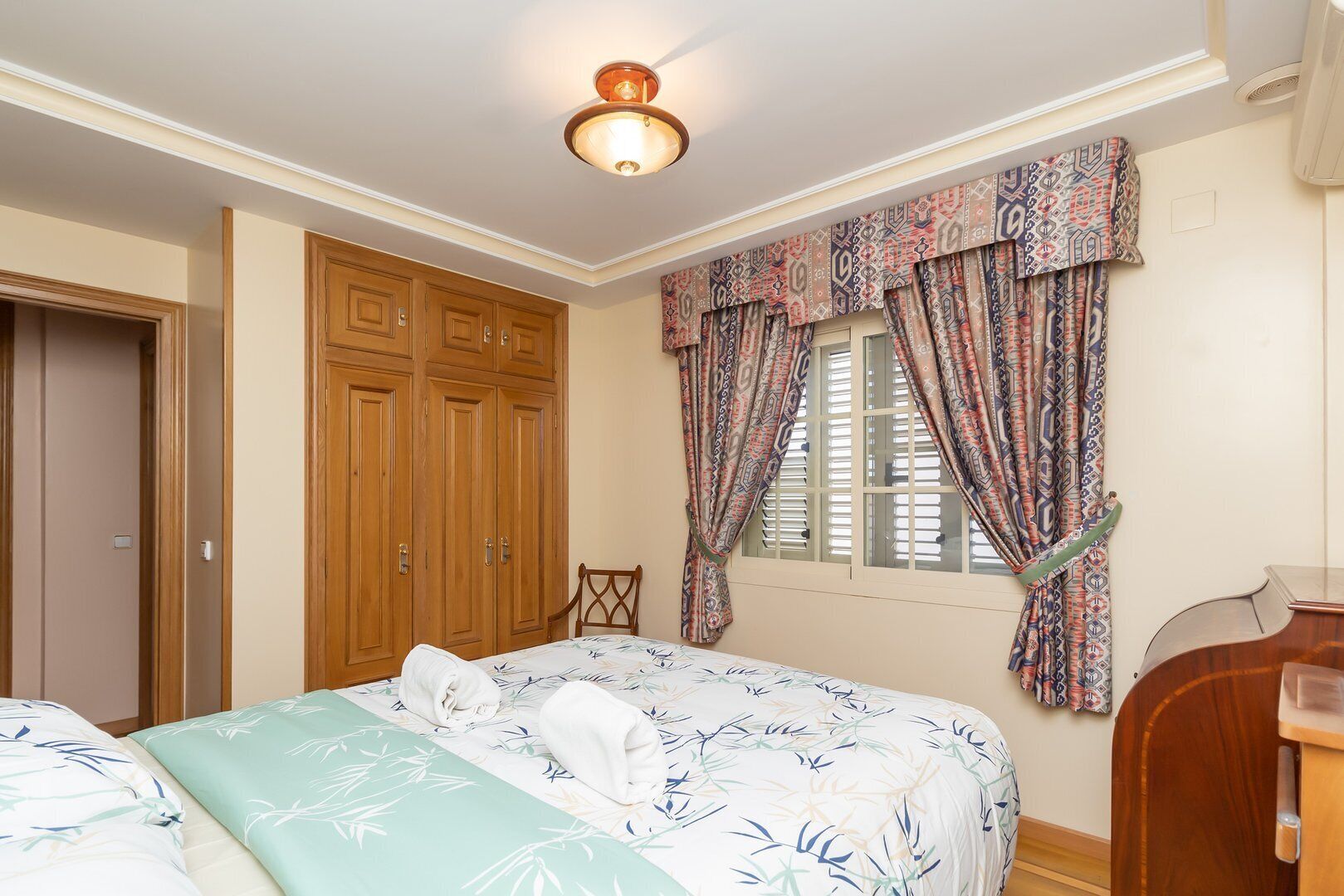 A bedroom with a bed, a wooden wardrobe, a chair, and a window with patterned curtains.