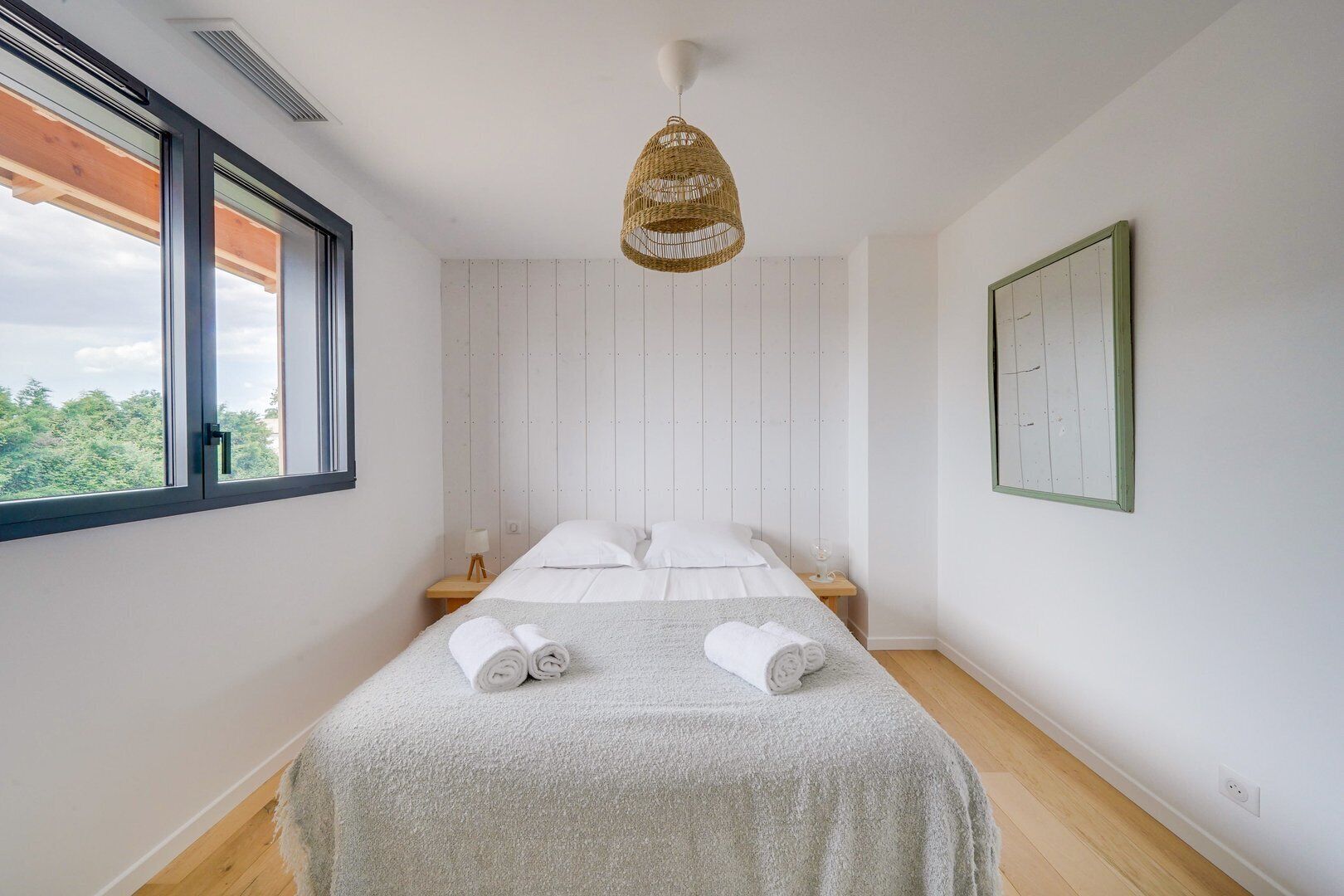 Een slaapkamer met een bed, een raam, een hanglamp en een aan de muur bevestigd fotolijstje.