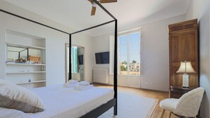 Een slaapkamer met een hemelbed, een stoel, een nachtkastje, een televisie, een raam en uitzicht op gebouwen.