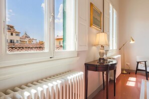 Ein Zimmer mit Heizkörper, Tisch mit Lampe, Stuhl und Fenster mit Blick auf Gebäude.