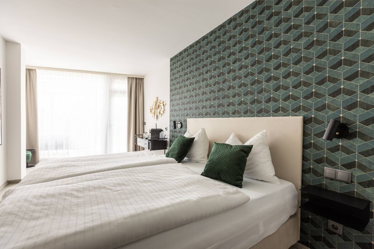 A hotel room with two beds, a geometric patterned wall, a bedside table, and a window with curtains.