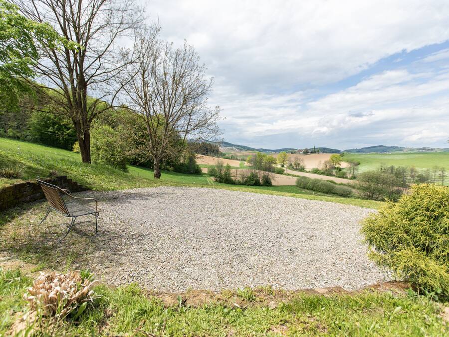 Un espace recouvert de gravier, avec un banc en métal, entouré de pelouse et d’arbres, offrant une vue sur un paysage rural.