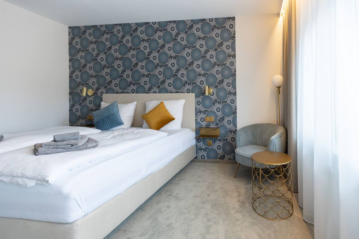 A neatly made bed with pillows, a bedside table, a chair, and a patterned wall.