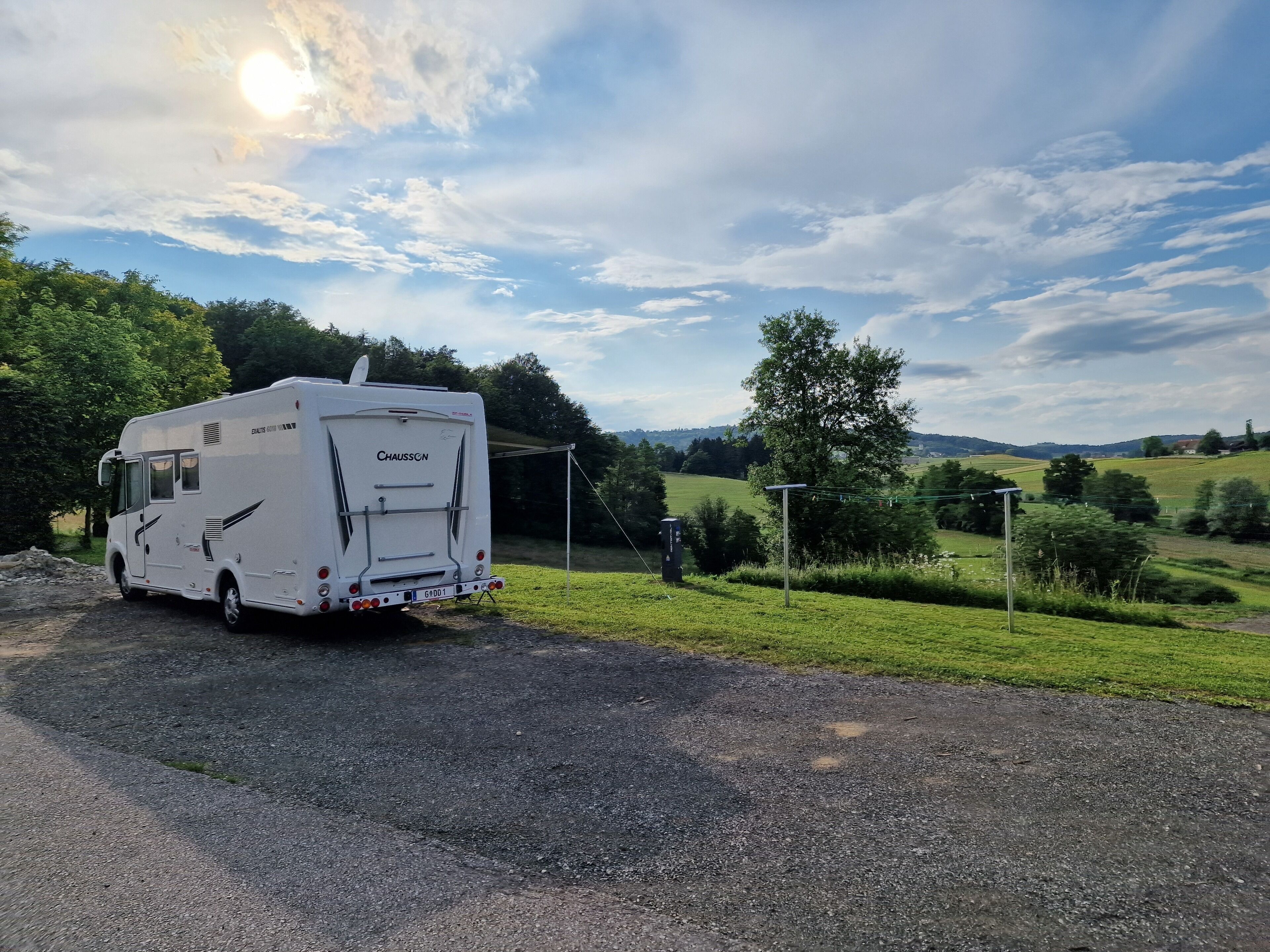 Ein weißer Wohnmobil auf einem Schotterweg mit schöner Aussicht auf grüne Felder und Bäume im Hintergrund.