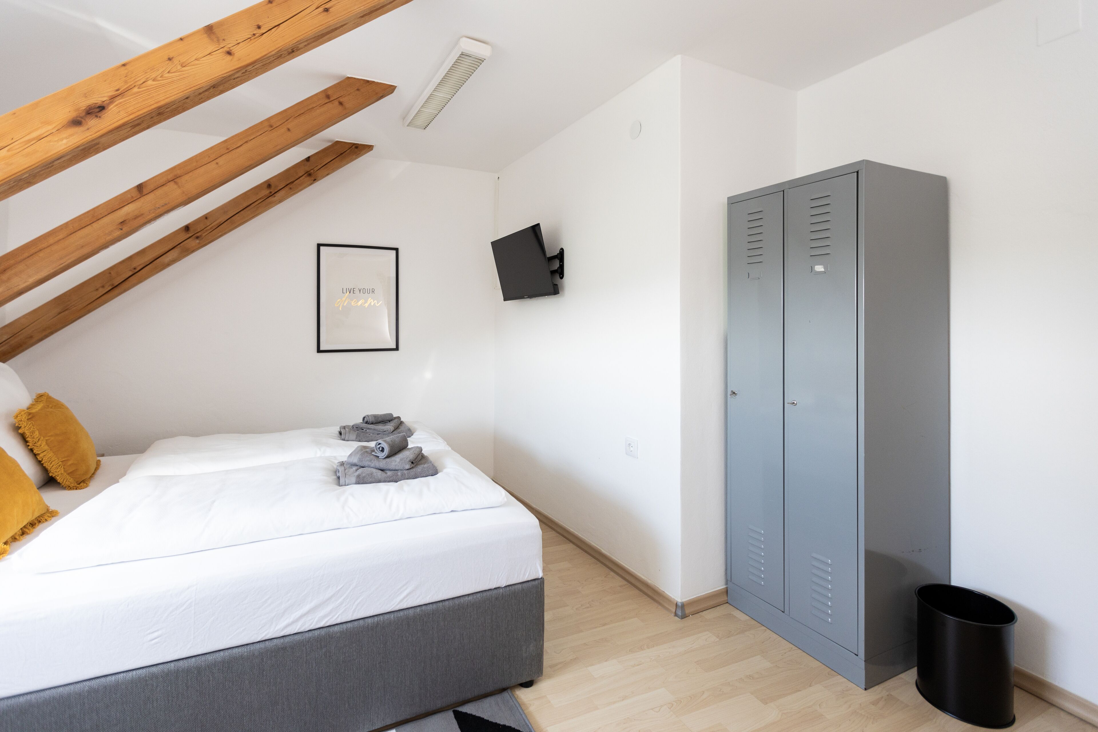 Un dormitorio con una cama, un armario gris, un cubo de basura negro y una cita motivacional enmarcada en la pared.