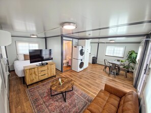 Espaço compacto para viver, com cama, TV, sofá, área de refeições e cozinha pequena com máquina de lavar.