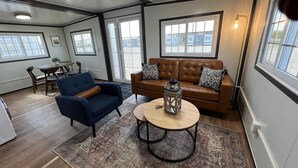 Un salon spacieux avec un canapé en cuir marron, un fauteuil bleu et une table basse ronde surmontée d’une lanterne. De grandes fenêtres laissent entrer la lumière naturelle et offrent une vue sur l’extérieur.