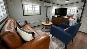 Un espace de vie compact comprenant un canapé en cuir marron, un fauteuil bleu, une petite table ronde, un téléviseur et une commode en bois.