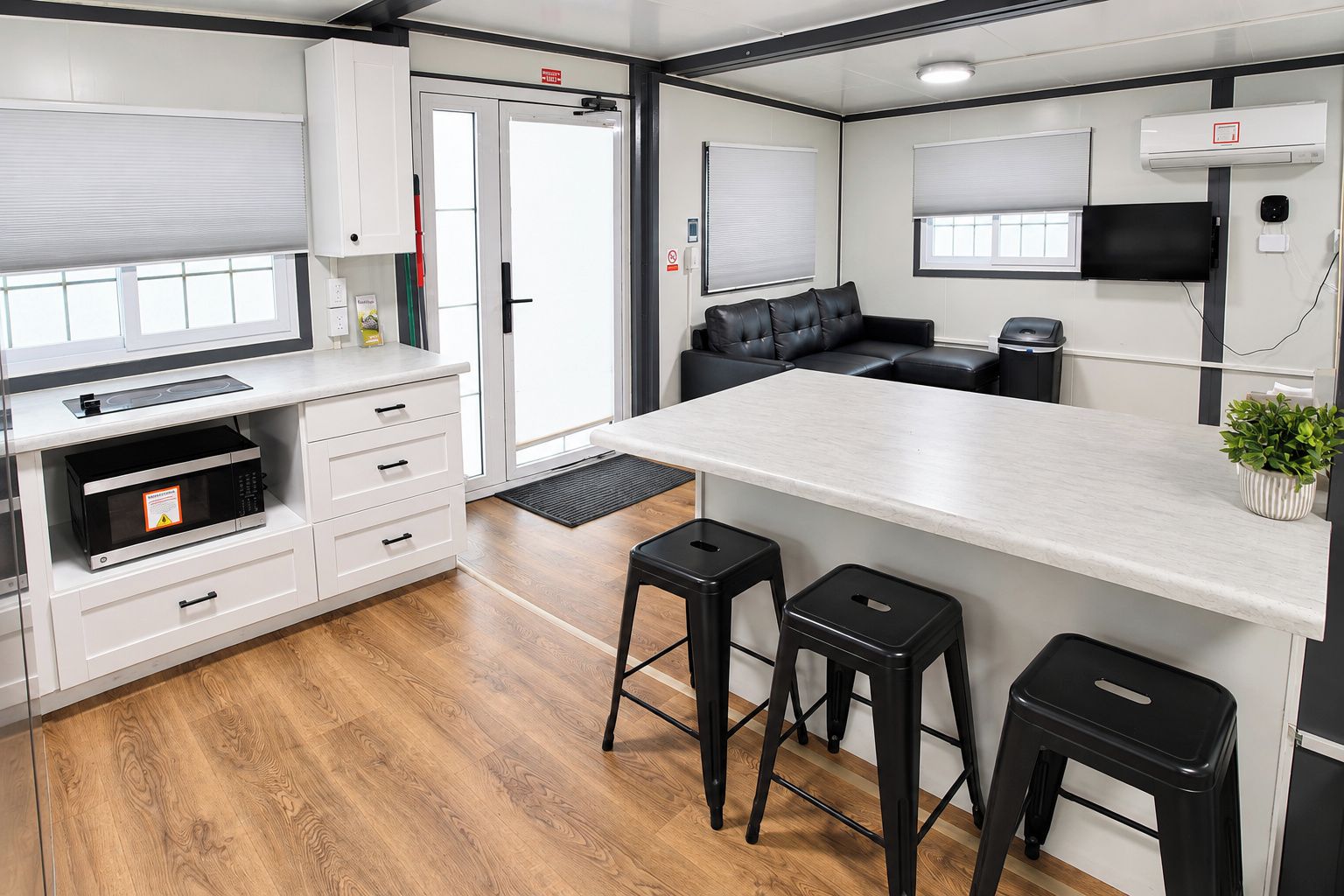 A compact kitchen with a microwave, a coffee machine, and a small refrigerator. There are two stools and a black leather sofa.