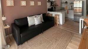 Ein modernes Wohnzimmer mit einem schwarzen Ledersofa, einem Deckenventilator und einer Küchenzeile inklusive Kühlschrank und Kaffeemaschine.