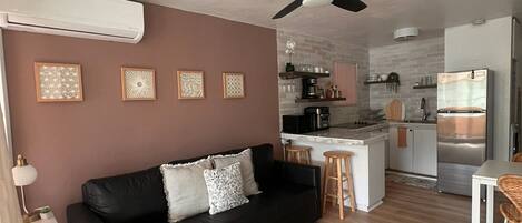 A modern living room with a black leather sofa, a ceiling fan, and a kitchen area with a refrigerator and a coffee maker.
