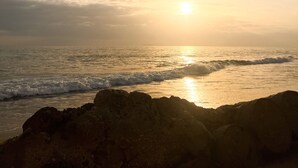 Eine Strandansicht mit untergehendem Sonnenuntergang über dem Meer, Wellen, die an das Ufer schlagen, und Felsformationen im Vordergrund.