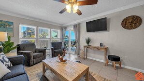 A living room with a ceiling fan, a TV mounted on the wall, and a view of the outdoors through large windows.