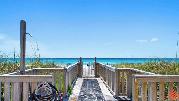 A wooden boardwalk leading to a beach with clear blue water and a grassy area.