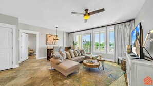 A spacious living room with a beige sofa, a coffee table, and a TV. There is a ceiling fan, a dining area with a table and chairs, and large windows with curtains. - The Grand in SanDestin Resort Luxury 1BR Condo (Miramar Beach)