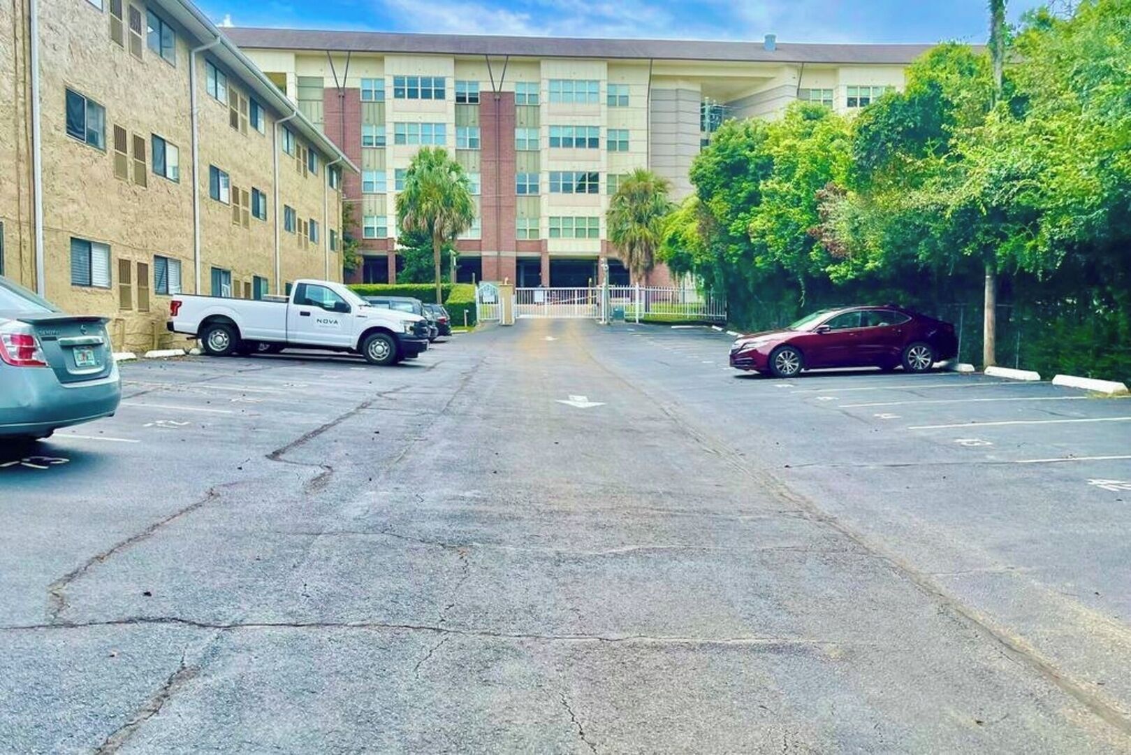 A parking lot with a white truck and a red car, a multi-story building with a flat roof, and trees along the side.