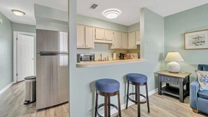 A compact kitchen with a refrigerator, microwave, and wooden cabinets. There are two bar stools and a small table with a lamp.