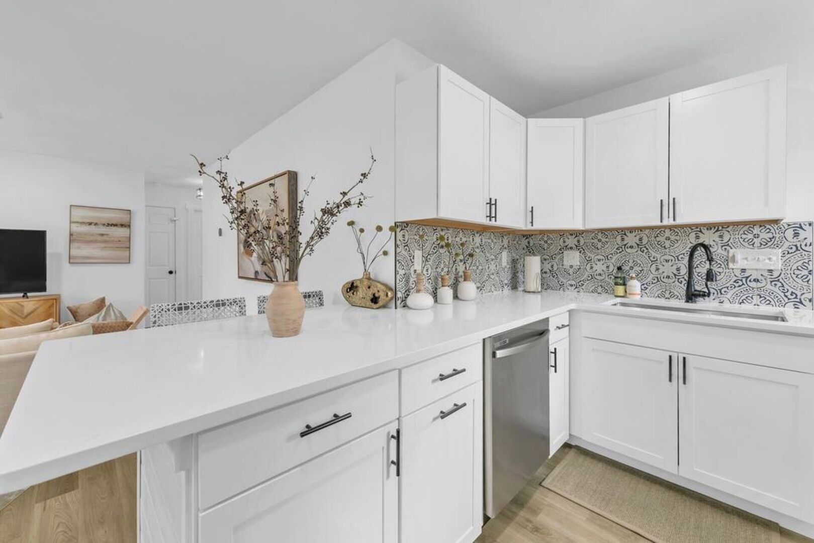 A modern kitchen with white cabinetry, a white countertop, and a backsplash with a black and white pattern. There are decorative vases and a framed picture on the countertop.