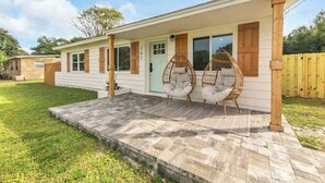 Una casa de un solo piso con un porche cubierto, dos sillas de mimbre y un patio de piedra.