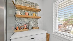 Cocina con armarios blancos, encimera blanca y salpicadero con diseño. Estantes de madera sobre la encimera contienen tazas y frascos.
